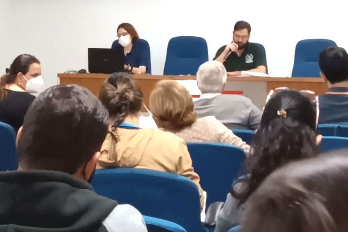 Na foto aparecem uma mulher e um homem, sentados atrás de uma mesa, e cabeças várias pessoas que estão de costas, sentadas, olhando para as duas pessoas que compõem a mesa de coordenação da reunião.