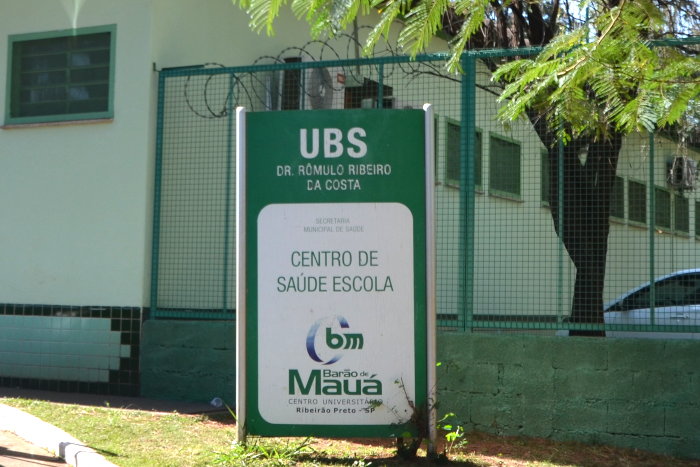 Na foto aparece uma placa com a inscrição "UBS Dr. Rômulo Ribeiro da Costa, Centro de Saúde Escola, Barão de Mauá - Centro Universitário, Ribeirão Preto-SP. No meio da placa aparece também a logomarca da faculdade, formada por um círculo cortado pelas letras bm, tudo na cor verde.