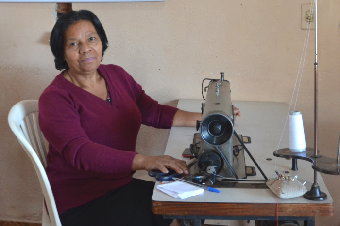 Na foto, uma mulher negra, com cabelos pretos curtos, blusa cor de vinho de mangas compridas e saia preta, está sentada em uma cadeira de plástico branca, diante de uma mesa que tem a máquina de costura industrial, com a mão direita sobre uma tesoura.