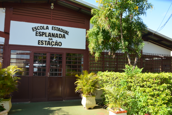 A foto é da frente da escola, mostrando a porta de entrada, com o nome "Escola Estadual Esplanada da Estação" escrito em letras grandes logo acima e mostrando as plantas que ficam dos dois lados da calçada que vai do portão da rua até a porta de entrada da casa.