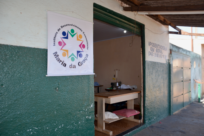 Foto da fachada da casa conhecida como "Salão da Anita", tendo ao centro uma porta aberta, com uma mesa do lado de dentro, que funciona como balcão de atendimento. Do lado esquerdo da porta, está o banner do Instituto de Desenvolvimento Comunitário Maria da Graça, com sua roda de bonequinhos coloridos de mãos dadas e no entorno da roda o nome da entidade. Do lado direito, aparece a o aviso "Aposentadorias em geral". A metade superior da parede é pintada de branco e a parte de baixo é verde. 