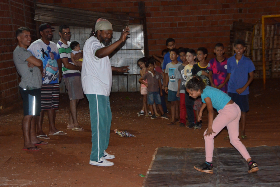 Na foto, um homem negro em pé, com camiseta branca e calça verde, tem seus braços levantados acenando para a menina que está sobre a lona, fazendo um dos movimentos ensinados pelo Mestre. Atrás, uma fila de outras crianças e adolescentes. Do lado esquerdo, três homens em pé observam a aula