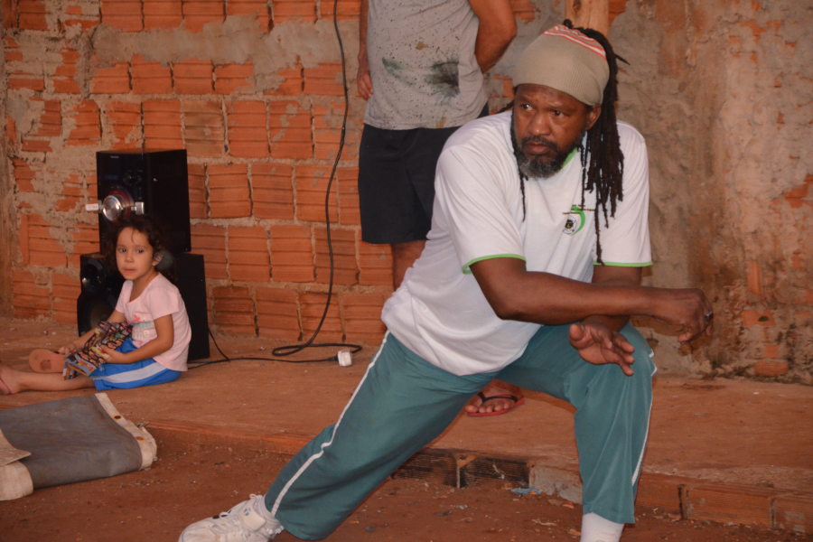 Na foto, um homem negro, de cabelos compridos com dreads e uma bandana cinza, camiseta branca e calça verde, tem uma das pernas esticadas, apoiando-se sobre a outra que está dobrada, na demonstração de um dos movimentos da capoeira. Ao fundo, uma criança sentada no chão. 