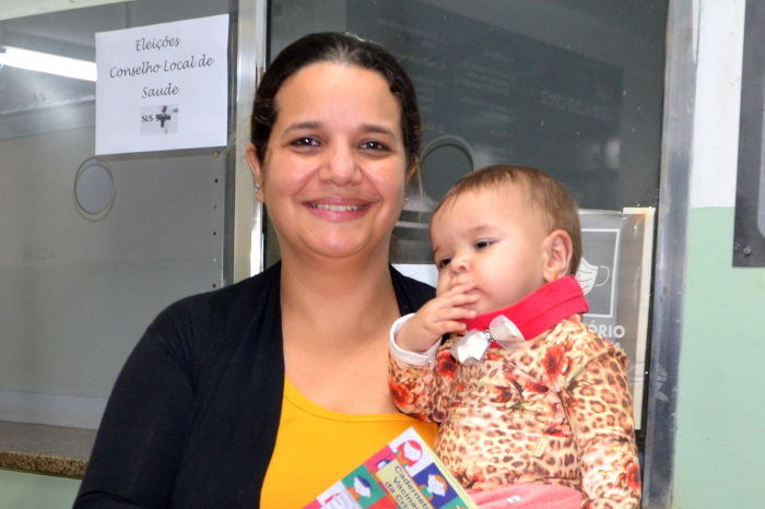 Na foto, uma mulher branca, com cabelos castanhos amarrados atrás da cabeça, sorridente, com uma criança nos braços