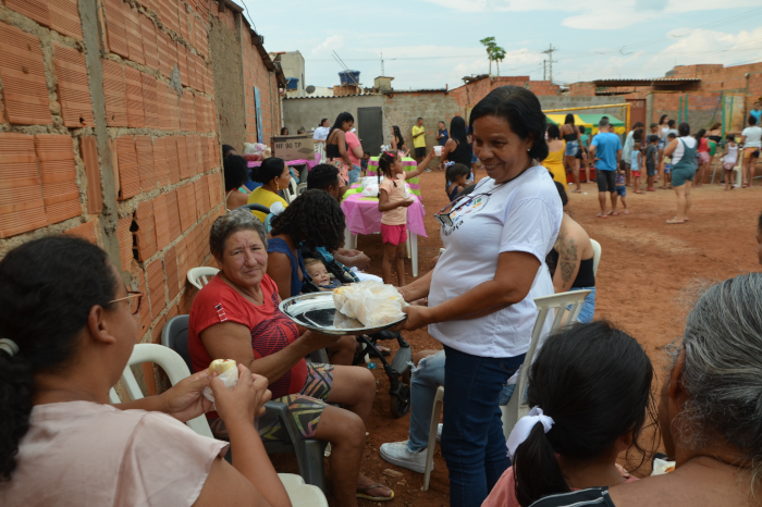 Na foto, uma mulher negra, vestida com camiseta branca onde se vê a logomarca do Instituto Maria da Graça, tem uma bandeja cheia de sanduíches na mão. Várias outras mulheres estão sentadas, algumas com sanduíche na mão. Atrás, aparecem muitas pessoas, homens mulheres e crianças