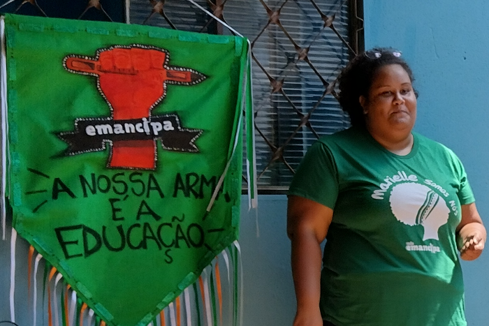 Na foto, um banner em formato de flâmula tem a imagem de um punho fechado segurando um lápis com a inscrição "Emancipa - A nossa arma é a educação". Ao lado da flâmula, uma mulher negra, com cabelos pretos e curtos, tem um óculos no alto da cabeça e está com uma camiseta verde onde se lê "Marielle somos nós" escrito acima da figura de uma cabeça representando a vereadora Marielle assassinada no Rio de Janeiro