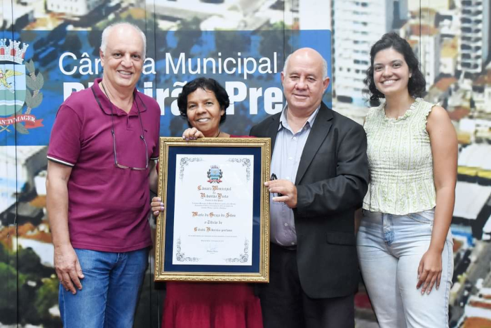 Na foto estão quatro pessoas, todas em pé: um homem branco meio careca, com camisa cor de vinho, ao lado da Anita, uma mulher negra, com cabelos curtos e pretos e vestido vermelho, que está segurando um quadro com vidro e moldura. Do seu lado direito está outro homem, meio careca e vestindo um terno preto e uma mulher também negra, com cabelos curtos e vestindo calça jeans e blusinha amarela clara