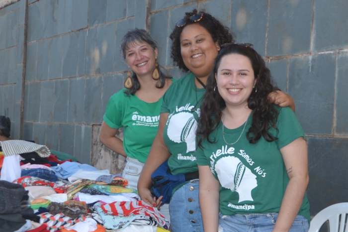 Na foto, três mulheres, todas de camiseta verde, ao lado de uma mesa com pilhas de roupas. Da esquerda para a direita, a primeira é branca, de cabelos com mechas claras; a segunda é negra e tem cabelos pretos e curtos; e a terceira é branca, com cabelos pretos cacheados e compridos 