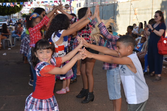 Na foto aparecem crianças e adolescentes dançando a quadrilha no momento em que fazem o túnel. Em duas filas, uma de frente para outra, todos estão com os braços levantados e as mãos cada uma tocando a do seu par que está na sua frente.  Em primeiro plano, uma menina com vestido colorido de festa junina tem seus dedos das duas mãos entrelaçados aos dedos das duas mãos de um menino que está na sua frente 