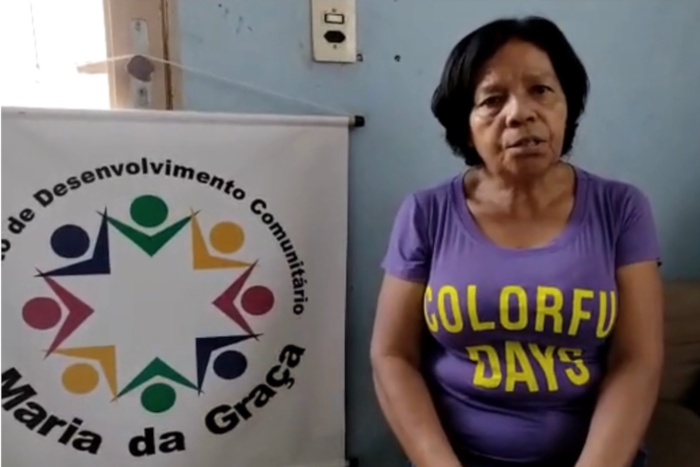 Na foto, uma mulher negra, de cabelos pretos e curtos, com uma camista lilás, tendo ao lado um banner com a logomarca do Instituto Maria da Graça