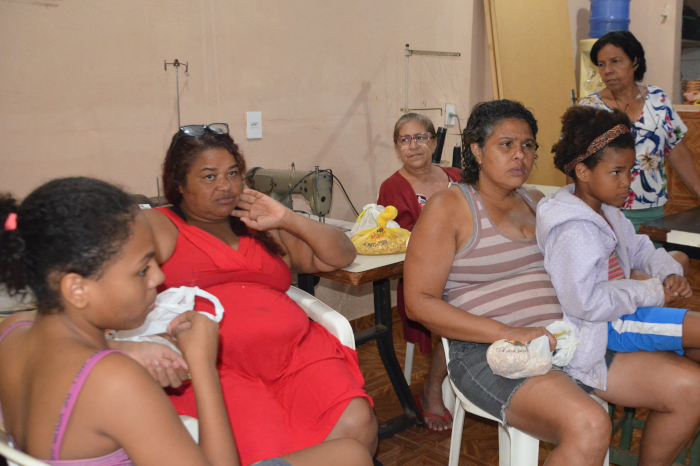 Na foto aparecem seis mulheres sentadas, uma com uma criança no colo, destacando-se uma mulher negra, com cabelos compridos, soltos, e vestido vermelho, que é representante do bairro no Conselho de Saúde