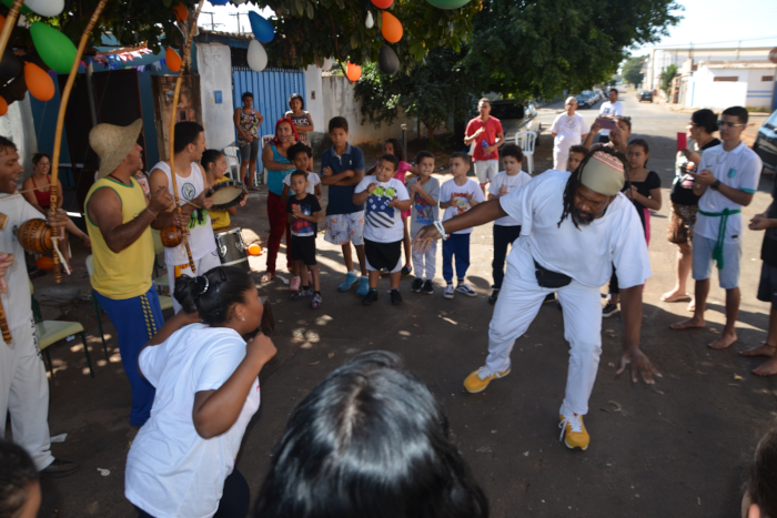 Na foto, um homem negro, com calça e camisa branca, joga capoeira com um menina também negra, com cabelo amarrado em coque atrás da cabela e vestida com camiseta branca. Eles estão no centro de uma roda formada por crianças, adolescentes e adultos, todos em pé, onde aparecem dois homens tocando berimbau e outro tocando pandeiro