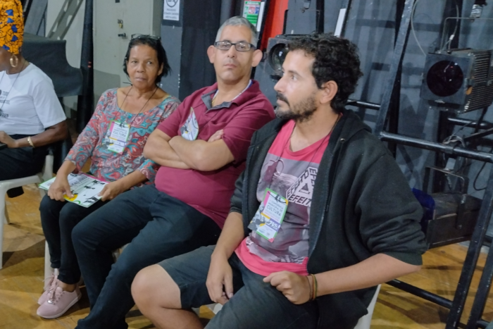 Na foto, três pessoas sentadas, todas com crachás da Conferência. Uma mulher negra, de cabelos curtos e pretos, com blusa estampada, um homem branco de cabelos curtos e óculos, com camisa vermelha e um outro homem, branco, com barba curta, está com moletom preto, bermuda preta e camiseta vermelha