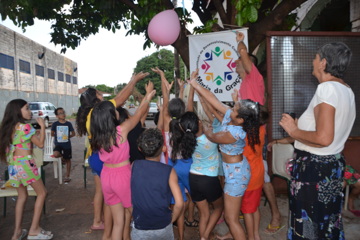 Na foto, várias crianças pulando com os braços levantados em direção a um grande balão cor de rosa à espera de que seja estourado para disputarem as balas que cairão. Ao fundo, o banner com a inscrição Instituto de Desenvolvimento Comunitário Maria da Graça em torno da logomarca do Instituto