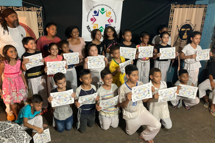 Na foto, 16 crianças segurando um papel com as duas mãos, sendo que a primeira fileira está ajoelhada no chão e na segunda todos estão em pé. Atrás da segunda fileira, aparecem o Mestre Caxambu, Anita da Silva e mais três jovens. Todos que estão segurando o certificado também estão com a corda cinza amarrada na cintura 