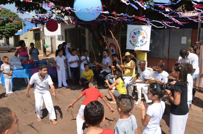 Na foto, várias crianças em pé, formando uma roda, junto com seis homens sentados, que tocam instrumentos como berimbau, pandeiro, agogô e tambor. A maioria está com roupas brancas, inclusive um dos meninos que aparece gingando no centro da roda em frente de outro menino que está com calça branca e camiseta vermelha