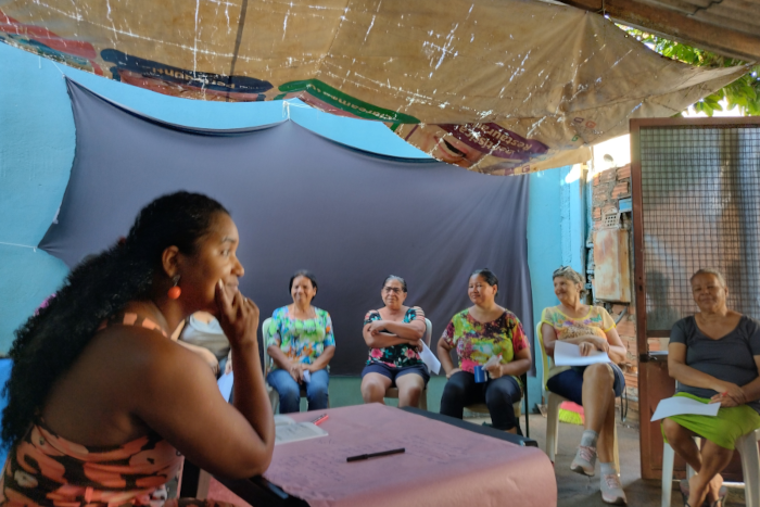 Do lado esquerdo, uma mulher negra, de cabelos compridos e pretos, está sentada, com o braço direito dobrado, a mão no queixo e olhando para várias outras mulheres também sentadas num semi círculo. À sua frente, aparecem cinco mulheres também sentadas, que olham para a a que está na frente