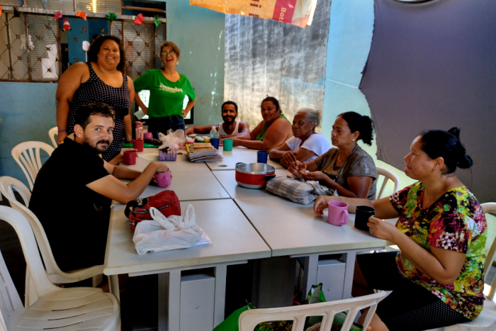 Na foto aparecem oito pessoas em torno de uma mesa, estando quatro mulheres e dois homens sentados e duas mulheres em pé.  O primeiro homem, sendado do lado esquerdo da imagem, é branco e tem cabelos e barbas pretas, estando vestido com camiseta preta. Em seguida aparece uma mulher negra, em pé, com vestido preto de bolinhas brancas. Depois está uma mulher branca, de cabelos loiros, vestida com camiseta verde estampada com a palavra Emancipa. Depois vem um homem negro, de cabelos e barba preta, que está com camiseta branca regata. Uma mulher negra, com blusa verde, outra mulher negra, de cabelos grizalhos, com blusa branca, outra mulher negra, com blusa cinza e uma outra mulher negra com blusa de estampa colorida. Todas essas mulheres sentadas têm os cabelos presos atrás da cabeça. Em cima da mesa há vários objetos, como uma panela, canecas e pequenas bolsas e sacolas