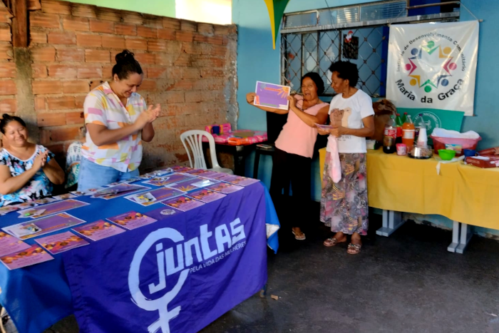 Na foto aparecem quatro mulheres. Duas estão batendo palmas, uma está segurando e mostrando o seu certificado, um papel colorido que ela segura com as duas mãos, uma de cada lado da folha. A outra mulher está em pé ao seu lado. Na frente delas, uma mesa tem vários certificados espalhados, junto com outros papeis. A bandeira do Coletivo Juntas, está aberta e pendente na lateral da mesa. A bandeira tem a  cor azul, com o símbolo da mulher e a palavra JUNTAS escrita em branco. Ao fundo uma outra mesa tem copos, garrafas de refrigerante e outros utensilios. Na parede atrás da mesa está pendurado um banner com a logomarca e a inscrição "Instituto de Desenvolvimento Comunitário Maria da Graça"