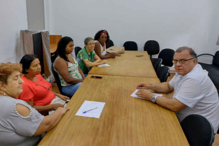 Cinco mulheres estao sentadas de um lado da mesa e um homem do outro. A primeira mulher que aparece na foto é branca, tem cabelos curtos e ruivos e está com uma blusa cinza claro. A segunda é negra, tem cabelos curtos, lisos e pretos, e está com uma blusa vermelha. A terceira também é negra, tem os cabelos encaracolados na altura do ombro e está com uma blusa branca, de alças, com estampas verdes. A quarta mulher é branca, usa óculos, tem cabelos grisalhos presos atrás da cabeça e está com uma blusa verde de estampas brancas. E a quinta mulher é negra, também usa óculos, tem cabelos pretos encaracolados e presos com um lenço vermelho, estando com uma blusa branca. O homem é branco, está usando óculos, tem cabelos curtos e grisalhos e veste uma camisa branca. As mulheres estão olhando para ele, e ele está olhando para a câmera na hora da foto.