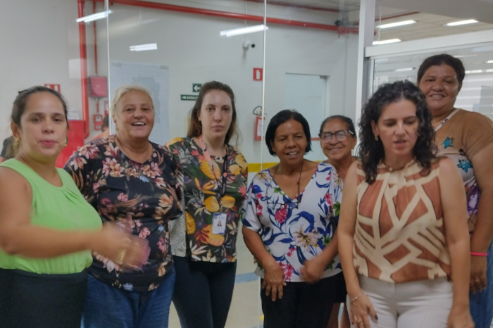 Na foto aparecem sete mulheres, todas em pé, umas ao lado das outras. Da esquerda para a direita, a primeira é branca, tem cabelos pretos amarrados atrás da cabeça e está usando uma blusa verde claro. A segunda também é branca, tem os cabelos brancos curtos e está usando uma blusa preta com flores coloridas. A terceira também é branca, com cabelos castanhos escuros compridos e soltos, está usando uma blusa de fundo preto com desenhos coloridos. A quarta é negra, com cabelos pretos e curtos e está usando uma blusa de fundo branco com desenhos coloridos. A quinta também é negra, tem os cabelos castanhos amarrados atrás da cabeça e usa óculos. A sexta é branca, tem os cabelos pretos e encaracolados soltos na altura dos ombros e está usando uam blusa com estampa marrom. E a quinta é negra, tem os cabelos amarrados atrás da cabeça e está usando uma blusa com desenhos coloridos.